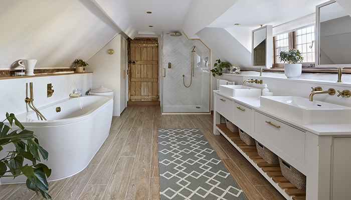 Simon Taylor Furniture combined a white handpainted bespoke vanity, white herringbone tiles, white basins, bath and sanitaryware with natural oak accents and timber-look porcelain tiles to create a soft spa-like environment in this en suite bath and shower room. To give it a luxury feel, they included brushed brass handles, wall-mounted taps and light fittings. They also maximised the natural daylight in the room by making swivel mirrors on brass poles above the his and hers basins